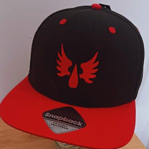 Könnte beinhalten: Eine schwarze Snapback-Cap mit einem roten, gestickten Flügelmotiv auf der Vorderseite. Die Cap hat einen flachen roten Schirm mit einem schwarz-silbernen Aufkleber, auf dem Snapback Original Headwear steht. Rote Ösen und ein roter Knopf oben.