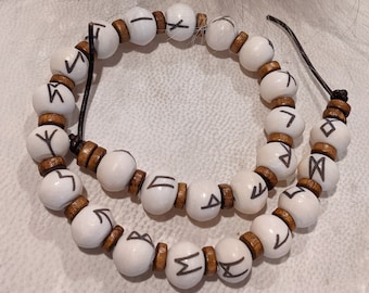 Op de afbeelding: Een ketting gemaakt van witte, ronde kralen met zwarte rune-symbolen en kleine bruine houten afstandhouders. De ketting is geregen op een zwart koord en wordt gepresenteerd op een wit oppervlak.