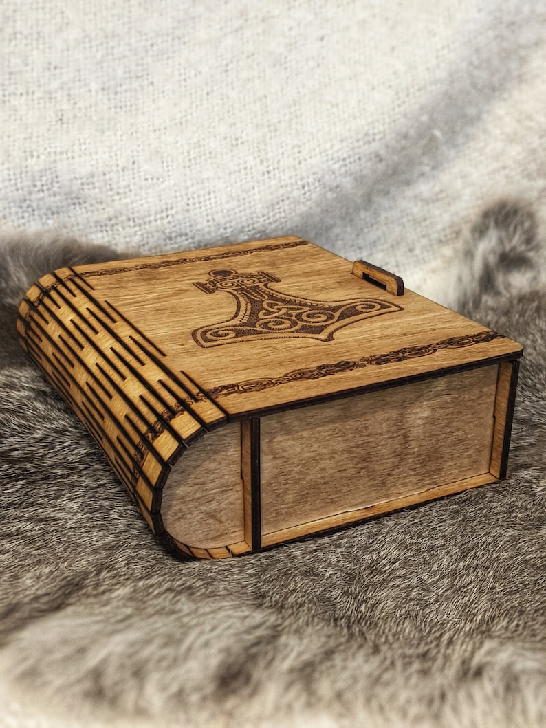 Op de afbeelding: Een houten doos met een scharnierend deksel, met een lasergegraveerd ontwerp van een hamer met ingewikkelde patronen. De doos is gemaakt van lichtbruin hout en heeft een natuurlijke afwerking.