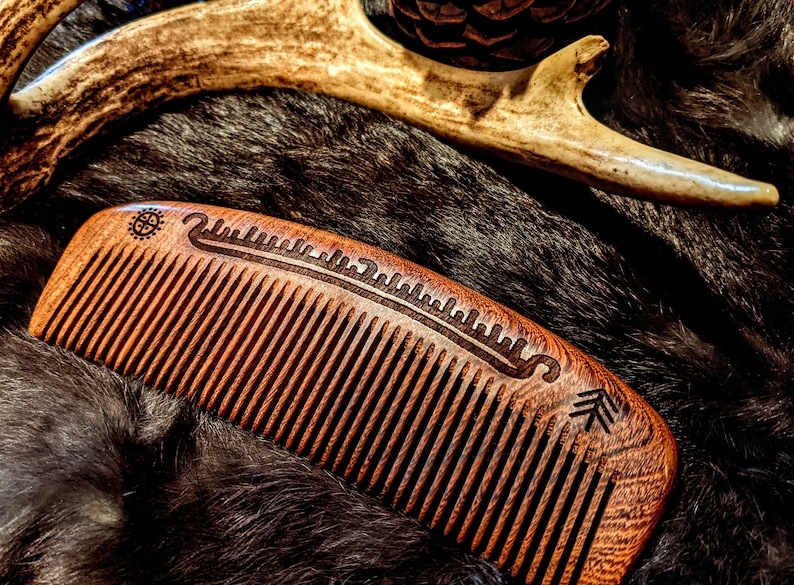 May include: A wooden comb with a carved design on the back. The comb is brown and has a natural finish. It is lying on a brown fur surface.