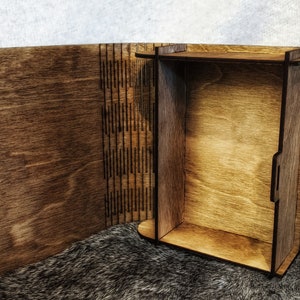 Op de afbeelding: Een houten doos met een scharnierend deksel dat opengaat om een ​​lege binnenkant te onthullen. De doos is gemaakt van donkerbruin hout en heeft een natuurlijke houten afwerking.