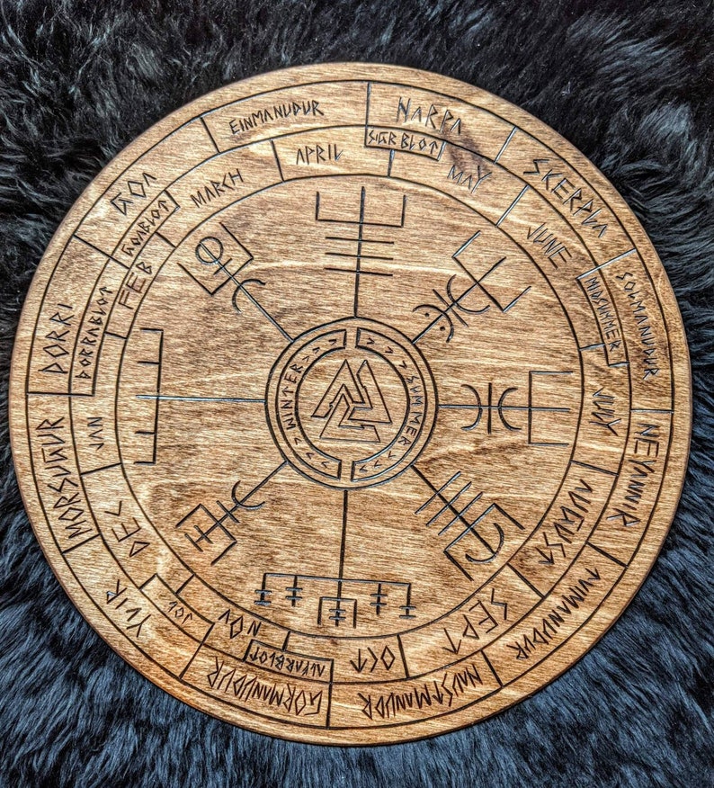 May include: A wooden circular calendar with Norse runes and symbols. The calendar features the months of the year in Old Norse, including Einmanudur, Narra, Skerrpla, and Solmanudur. The center of the calendar features a Vegvisir symbol, a Norse directional symbol.