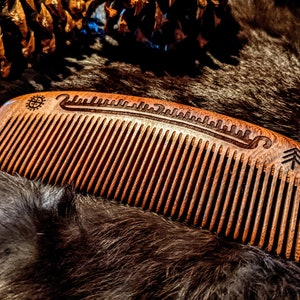 May include: A wooden comb with a carved design on the back. The design includes a tree, a wave, and a symbol. The comb is brown and has a natural finish.