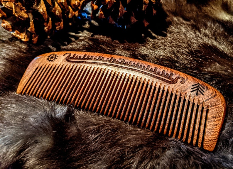 May include: A wooden comb with a carved design on the back. The design includes a tree, a wave, and a symbol. The comb is brown and has a natural finish.
