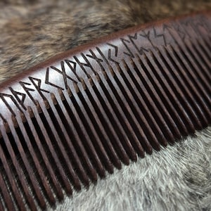 May include: A dark brown wooden comb with a row of teeth and runes carved into the top. The comb is lying on a light brown furry surface.