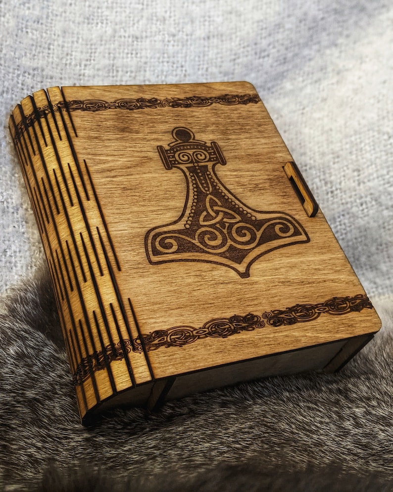 Op de afbeelding: Een houten doos met een scharnierend deksel en een gesneden ontwerp van een hamer met een knoopwerkpatroon. De doos is gemaakt van lichtbruin hout en heeft een donkerbruine vlek.
