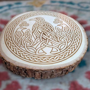 May include: A circular wooden slice featuring a laser-engraved design of three ravens surrounded by intricate Celtic knotwork patterns. The natural bark edge remains visible around the circumference of the light-colored wood surface.