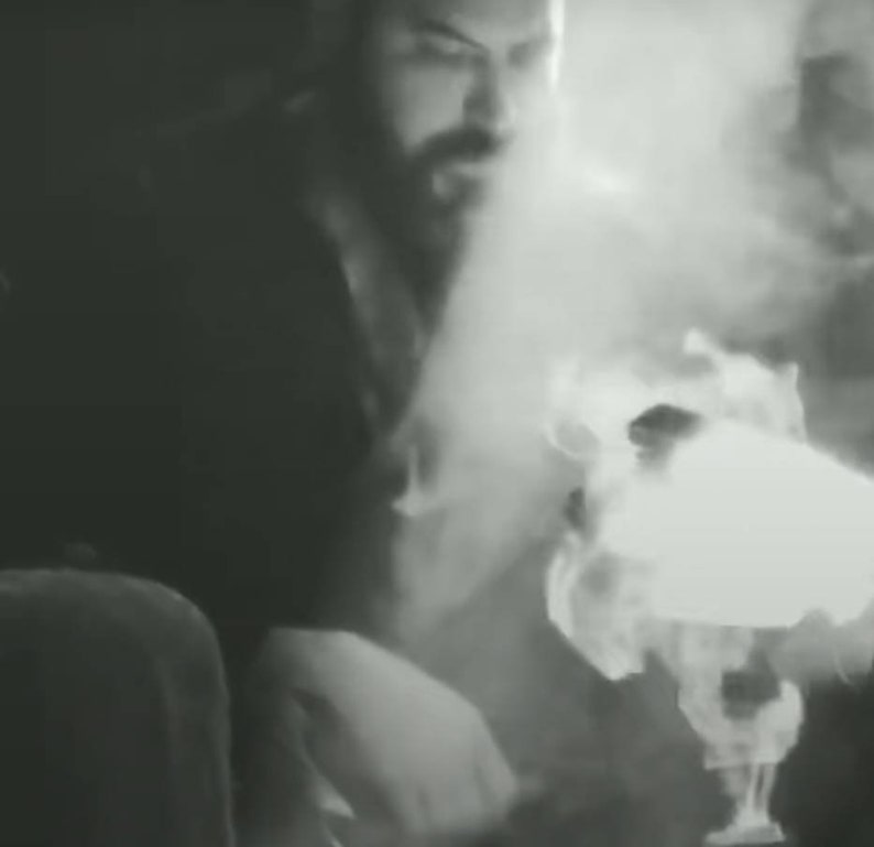May include: A close-up of a person's face with a beard, obscured by smoke or steam. The person is wearing a dark shirt and is looking at something off-camera.