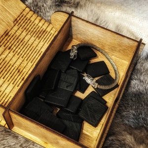 Op de afbeelding: Een houten doos met een scharnierend deksel, gevuld met zwarte houten runen en een zilveren armband met wolfshoofd accenten. De doos heeft een decoratief patroon op het deksel.