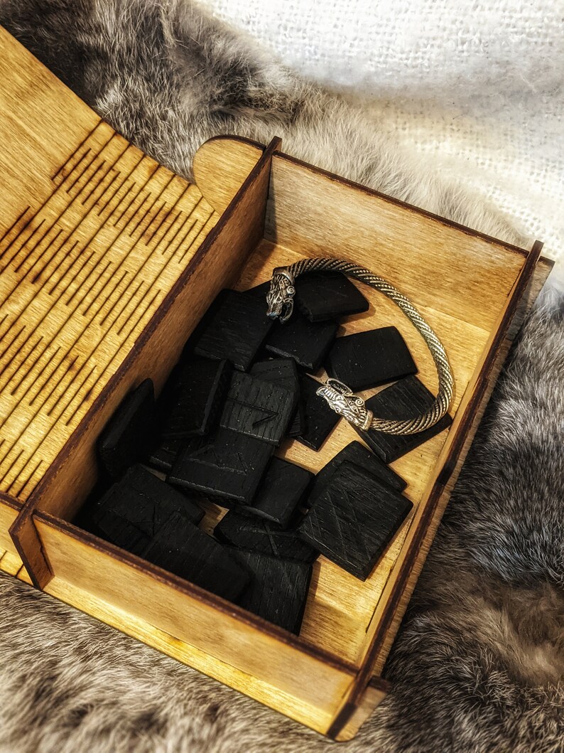 Op de afbeelding: Een houten doos met een scharnierend deksel, gevuld met zwarte houten runen en een zilveren armband met wolfshoofd accenten. De doos heeft een decoratief patroon op het deksel.