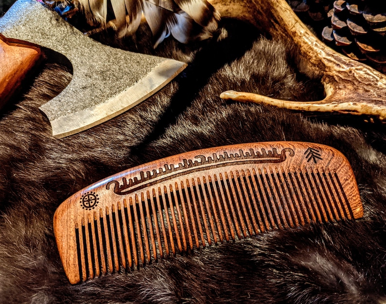 May include: A wooden comb with a carved design of a boat and a tree. The comb is brown and has a natural wood finish. The comb is lying on a brown fur surface.