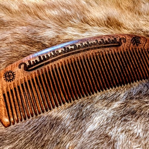 May include: A wooden comb with a carved design of a boat and a compass. The comb is brown and has a natural finish.