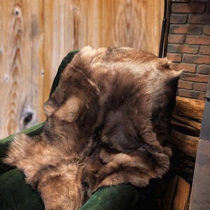 May include: A brown fur throw draped over a dark green velvet armchair. The throw has varying shades of brown and a soft, textured appearance. The background includes a wooden wall, a brick wall, and a lamp.