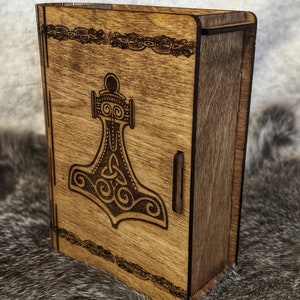 Op de afbeelding: Een houten doos met een scharnierend deksel, met een lasergegraveerd ontwerp van een gestileerde hamer met een Keltisch knoopmotief. De doos heeft een donkerbruine houtkleur en een decoratieve rand.