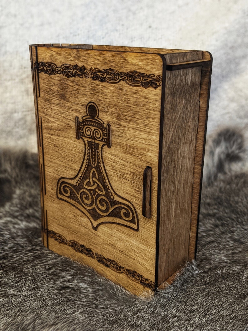 Op de afbeelding: Een houten doos met een scharnierend deksel, met een lasergegraveerd ontwerp van een gestileerde hamer met een Keltisch knoopmotief. De doos heeft een donkerbruine houtkleur en een decoratieve rand.