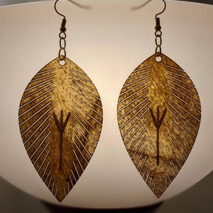 May include: A pair of wooden leaf-shaped earrings with a brown wood grain finish. Each earring features a small, engraved symbol in the center.