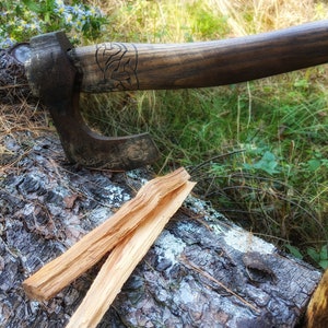 May include: A hand-forged axe with a wooden handle and a carved design. The axe is resting on a log with two pieces of kindling wood.