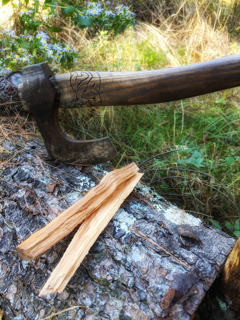 May include: A hand-forged axe with a wooden handle and a carved design. The axe is resting on a log with two pieces of kindling wood.