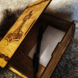 Op de afbeelding: Een houten doos met een scharnierend deksel, versierd met een lasergegraveerd ontwerp van een hamer. De doos is open en laat een gelinieerd notitieblok en een zwarte pen zien.