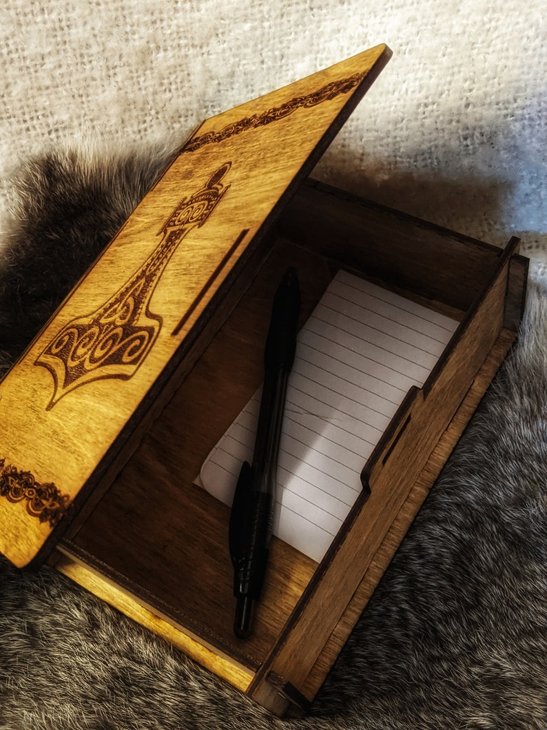 Op de afbeelding: Een houten doos met een scharnierend deksel, versierd met een lasergegraveerd ontwerp van een hamer. De doos is open en laat een gelinieerd notitieblok en een zwarte pen zien.