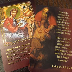 Może przedstawiać: Dwie karty z obrazami religijnymi. Lewa karta przedstawia obraz św. Łukasza piszącego w książce. Prawa karta przedstawia obraz mężczyzny obejmującego innego mężczyznę. Tekst na prawej karcie brzmi: "Szybko, przynieś najpiękniejszą szatę i odzież go w nią; włóż mu pierścień na palec i sandały na stopy. Ten syn mój był martwy, a ożył; był zagubiony i odnaleziony." - Łukasz 15: 22 & 24