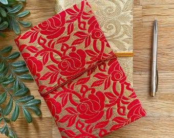 May include: Two notebooks with floral patterns. One notebook has a red and gold rose design, secured with a red cord. The other notebook is gold with a subtle floral pattern. A silver pen rests beside them.