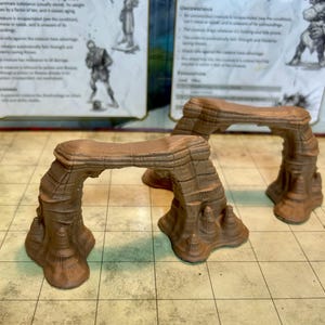 May include: Two brown, 3D printed, miniature stone archways. The arches are designed to be used as terrain pieces for tabletop games.