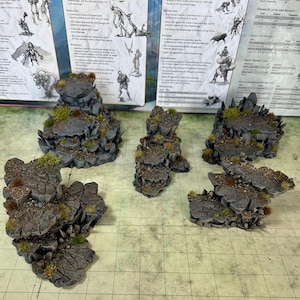 May include: Miniature rocky terrain pieces for tabletop gaming. The gray, textured outcrops feature patches of green and brown vegetation. The set includes various sizes and shapes, designed to create diverse landscapes for role-playing games and similar activities.
