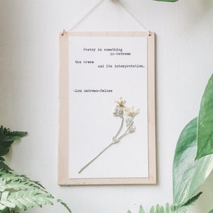 May include: A wooden sign with a white background and a quote about poetry by Lou Andreas-Salome. The quote reads "Poetry is something in-between the dream and its interpretation." A pressed flower is displayed on the right side of the sign.