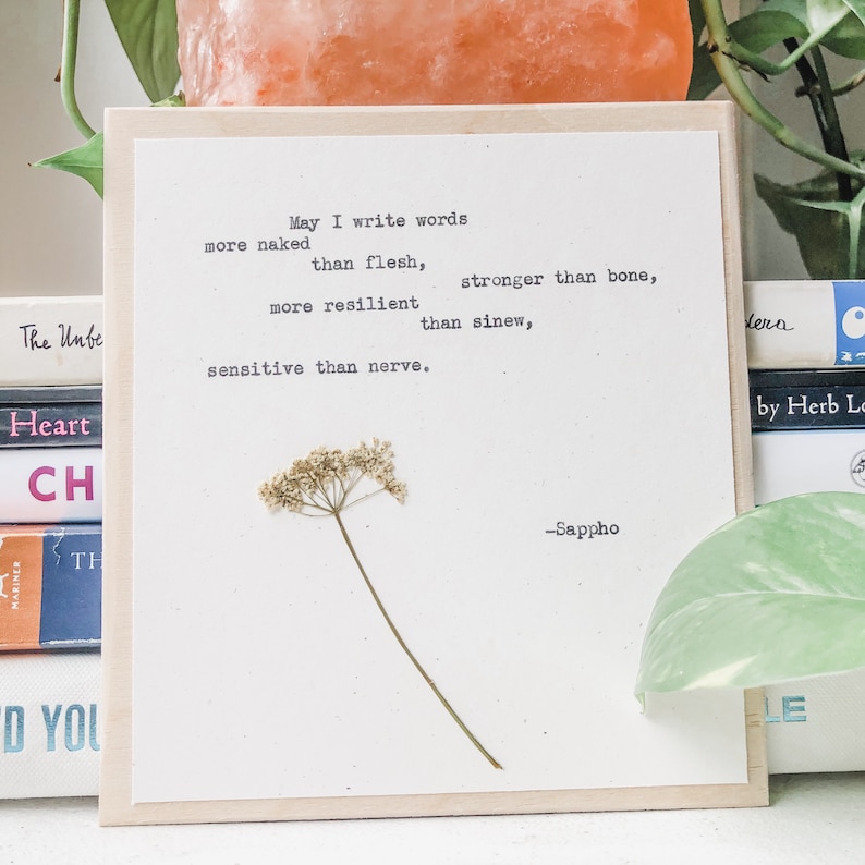 May include: A framed artwork featuring a pressed flower and a quote by Sappho. The quote, printed in black, reads "May I write words more naked than flesh, stronger than bone, more resilient than sinew, sensitive than nerve." The frame is light wood.