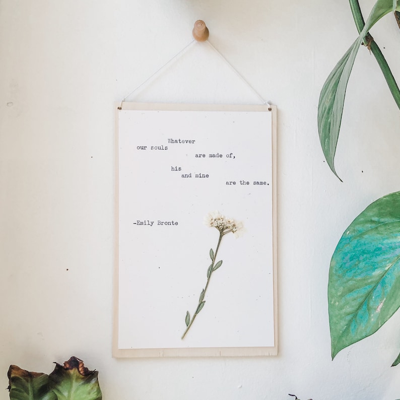 K&ouml;nnte beinhalten: Ein wei&szlig;er gerahmter Druck mit einem Zitat von Emily Bront&euml;: "Woraus auch immer unsere Seelen gemacht sind, seine und meine sind gleich." Eine kleine wei&szlig;e Blume ist in das Papier gepresst.
