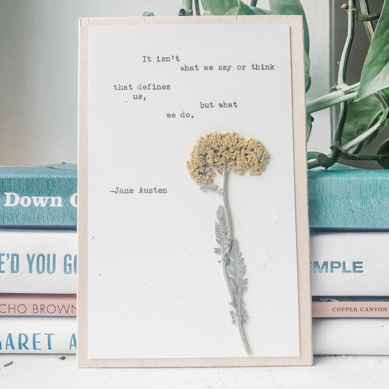 Puede incluir: Un letrero de madera blanco con una flor amarilla prensada y una cita de Jane Austen: "No es lo que decimos o pensamos lo que nos define, sino lo que hacemos."