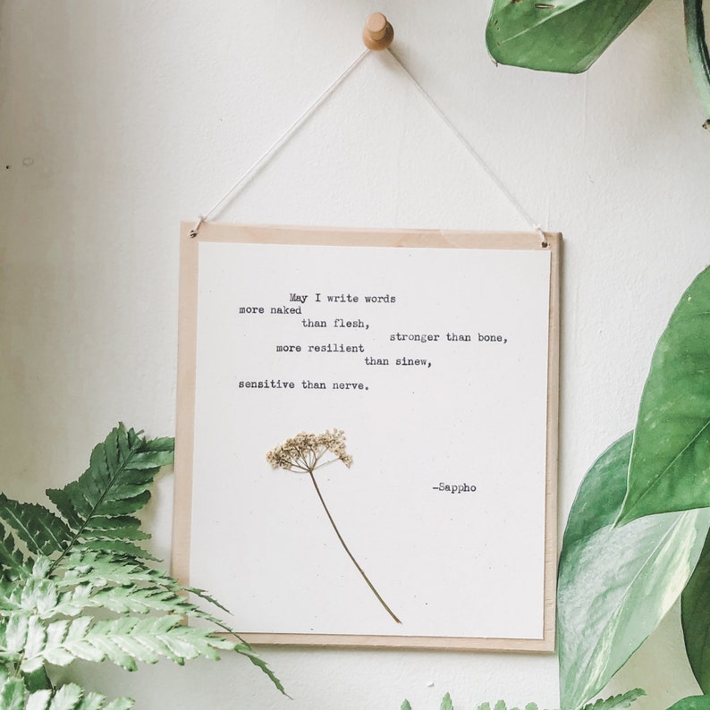 May include: A framed print with the quote "May I write words..." by Sappho. The print features a dried flower and is suspended by a string from a wooden peg. Green foliage surrounds the print, creating a natural aesthetic.
