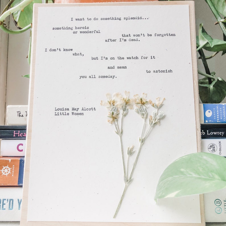 May include: A white card with a quote from Louisa May Alcott's Little Women, "I want to do something splendid... something heroic or wonderful that won't be forgotten after I'm dead. I don't know what, but I'm on the watch for it and mean to astonish you all someday."  The card has dried white flowers pressed on it.