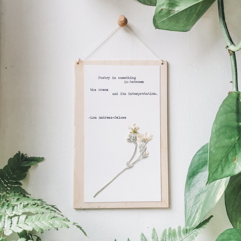 May include: A wooden sign with a white background and a quote about poetry by Lou Andreas-Salome. The quote reads "Poetry is something in-between the dream and its interpretation." A pressed flower is displayed on the right side of the sign.