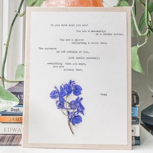 May include: A white card with a quote by Rumi printed on it. The quote reads: "Do you know what you are? You are a manuscript of a divine letter. You are a mirror reflecting a noble face. The universe is not outside of you. Look inside yourself; everything that you want, you are already that." A pressed purple flower is adhered to the card.