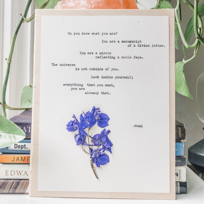 May include: A white card with a quote by Rumi printed on it. The quote reads: "Do you know what you are? You are a manuscript of a divine letter. You are a mirror reflecting a noble face. The universe is not outside of you. Look inside yourself; everything that you want, you are already that." A pressed purple flower is adhered to the card.