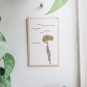 Puede incluir: Una impresi&oacute;n enmarcada en blanco con una cita de Jane Austen: "No es lo que decimos o pensamos lo que nos define, sino lo que hacemos." Una flor amarilla seca est&aacute; prensada en la impresi&oacute;n.