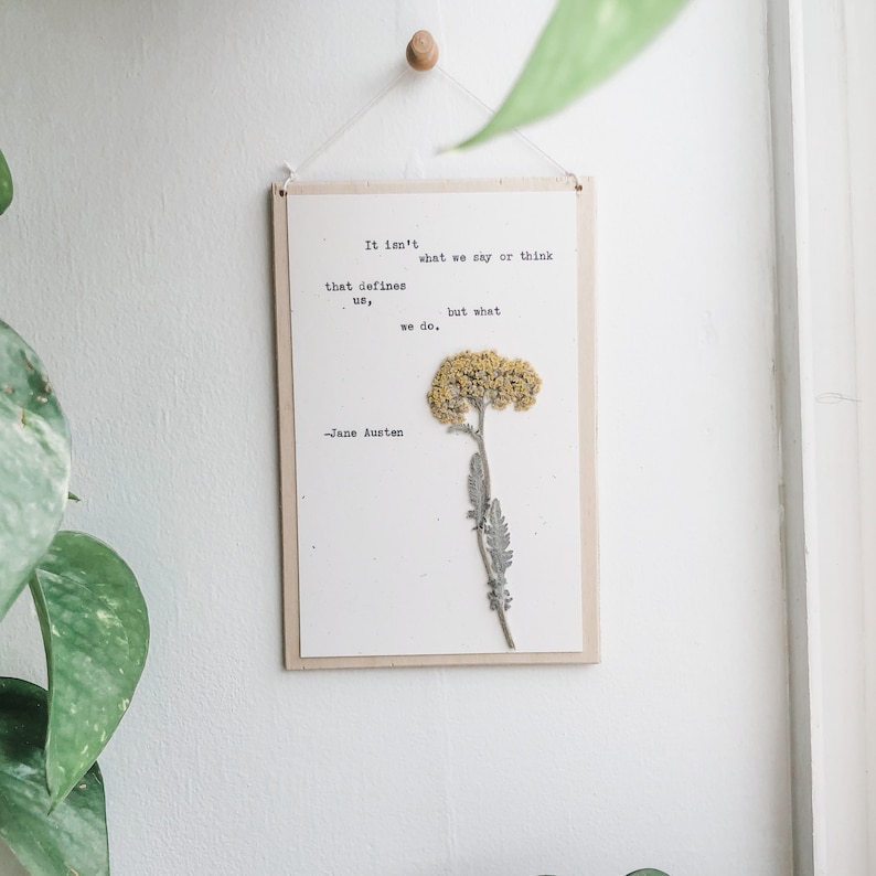 Puede incluir: Una impresi&oacute;n enmarcada en blanco con una cita de Jane Austen: "No es lo que decimos o pensamos lo que nos define, sino lo que hacemos." Una flor amarilla seca est&aacute; prensada en la impresi&oacute;n.