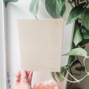 May include: A blank, light brown wooden sign with a smooth surface. The sign is held in a hand and is in front of a green plant.