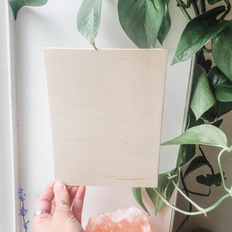 May include: A blank, light brown wooden sign with a smooth surface. The sign is held in a hand and is in front of a green plant.
