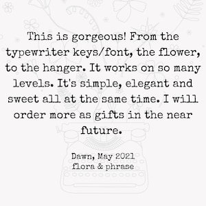 May include: Black and white image of a typewriter with a handwritten note that reads: "This is gorgeous! From the typewriter keys/font, the flower, to the hanger. It works on so many levels. It's simple, elegant and sweet all at the same time. I will order more as gifts in the near future. Dawn, May 2021 flora & phrase"