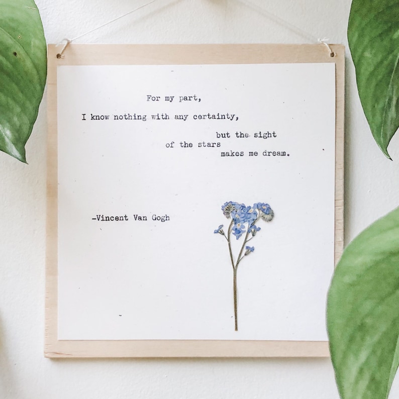 May include: A wooden sign with a white background and a quote by Vincent Van Gogh about the stars. The quote reads: "For my part, I know nothing with any certainty, but the sight of the stars makes me dream." A pressed blue flower is on the right side of the sign.