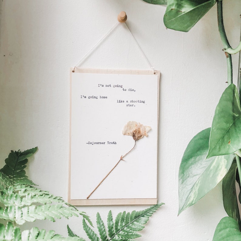 May include: A wooden sign with a quote by Sojourner Truth: "I'm not going to die, I'm going home like a shooting star." A dried flower is attached to the sign.