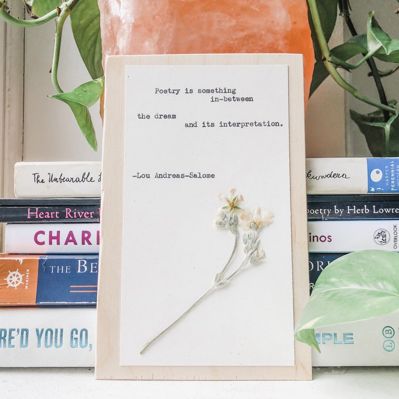May include: A white wooden sign with a quote about poetry by Lou Andreas-Salome. The quote reads "Poetry is something in-between the dream and its interpretation." A pressed flower is on the right side of the sign.