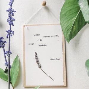 May include: A white wooden sign with a quote by the Dalai Lama: "Be kind whenever possible. It is always possible." The sign is hanging from a string and has a dried lavender sprig in the bottom right corner.