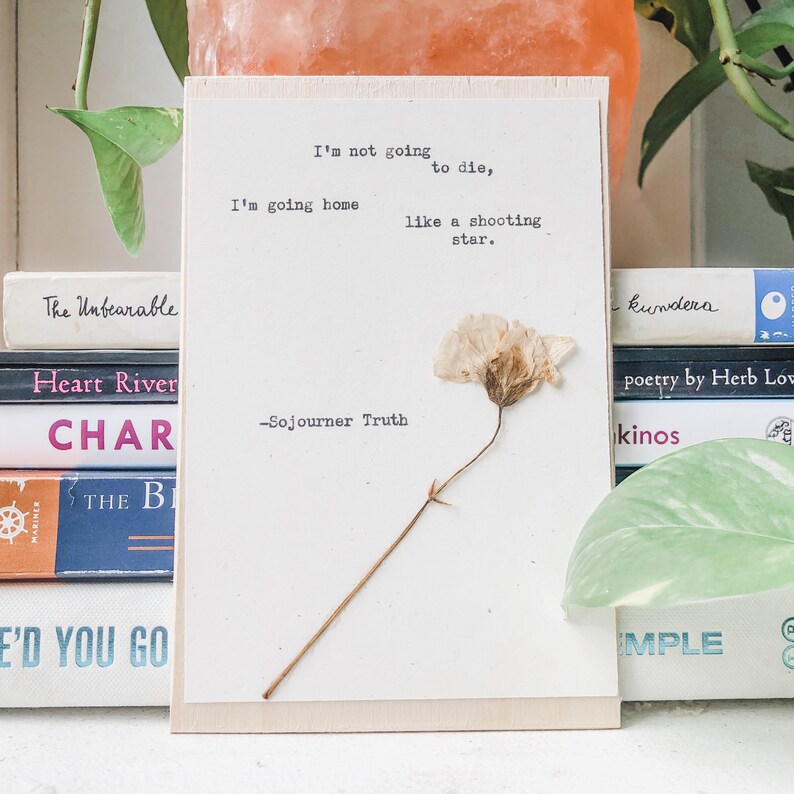 May include: A white wooden sign with a pressed flower and a quote by Sojourner Truth: "I'm not going to die, I'm going home like a shooting star."