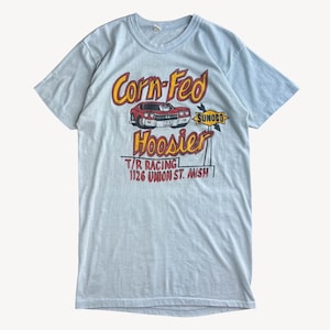 May include: Light blue t-shirt with a vintage graphic. The graphic features the words "Corn-Fed Hoosier" in yellow and red, a red car, and the text "T/R Racing! 1126 Union St. Mish". The shirt has a crew neck and short sleeves.
