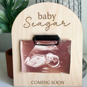 May include: A wooden sign with the text "baby Seagar coming soon" and a black clip holding an ultrasound image.