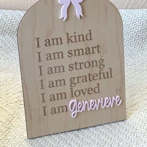 May include: A wooden sign with a pink bow at the top. The sign has the text "I am kind, I am smart, I am strong, I am grateful, I am loved, I am Genevieve" written on it.